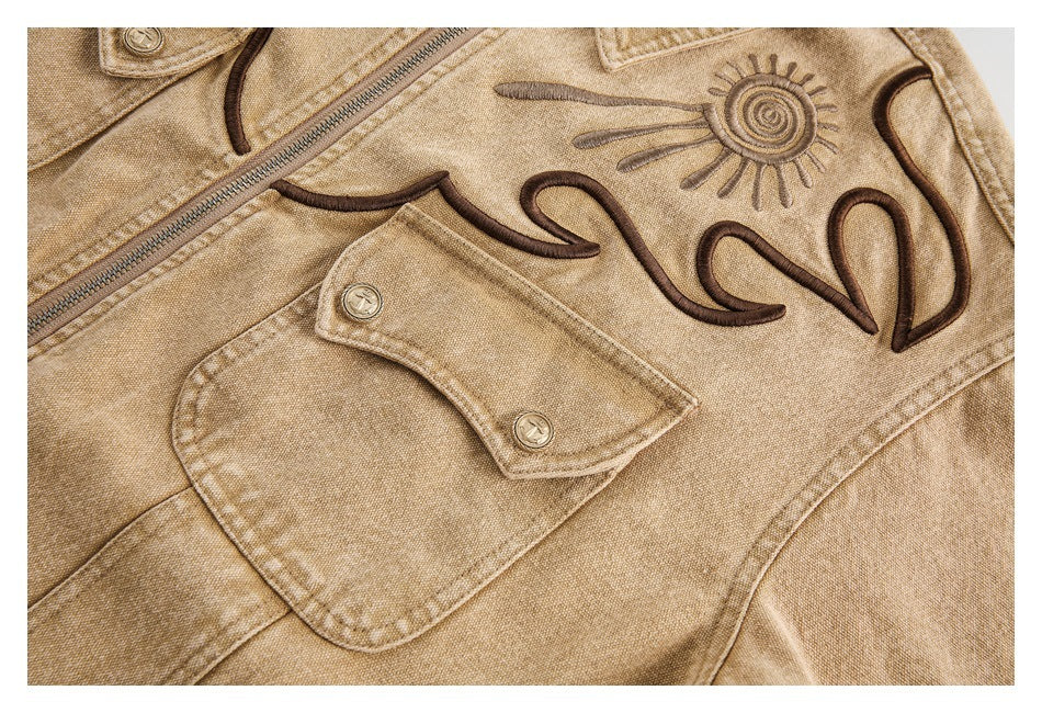 Close-up of a tan jacket's buttoned flap pocket and dark curved embroidery with a sun motif.