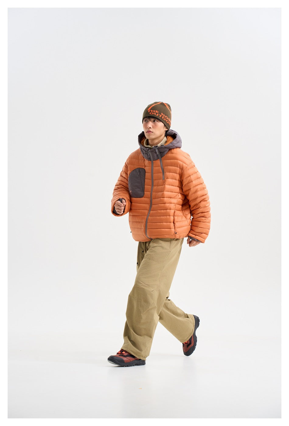 Model walking in an orange quilted hooded jacket with a gray chest pocket and olive trousers, shown from the front at an