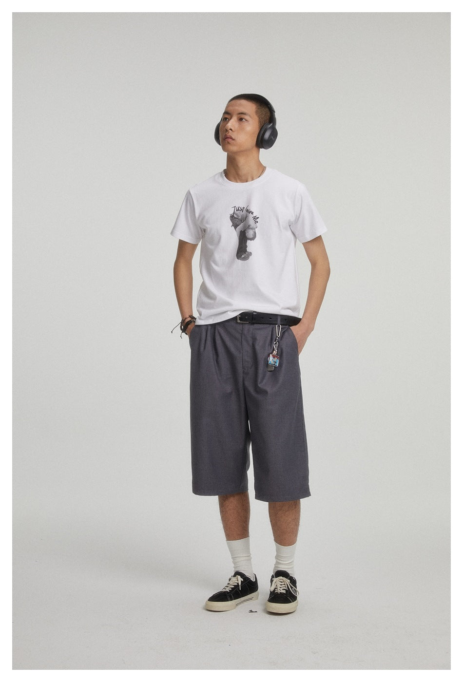 Model wearing a white graphic T-shirt and loose charcoal shorts, with headphones over his ears.