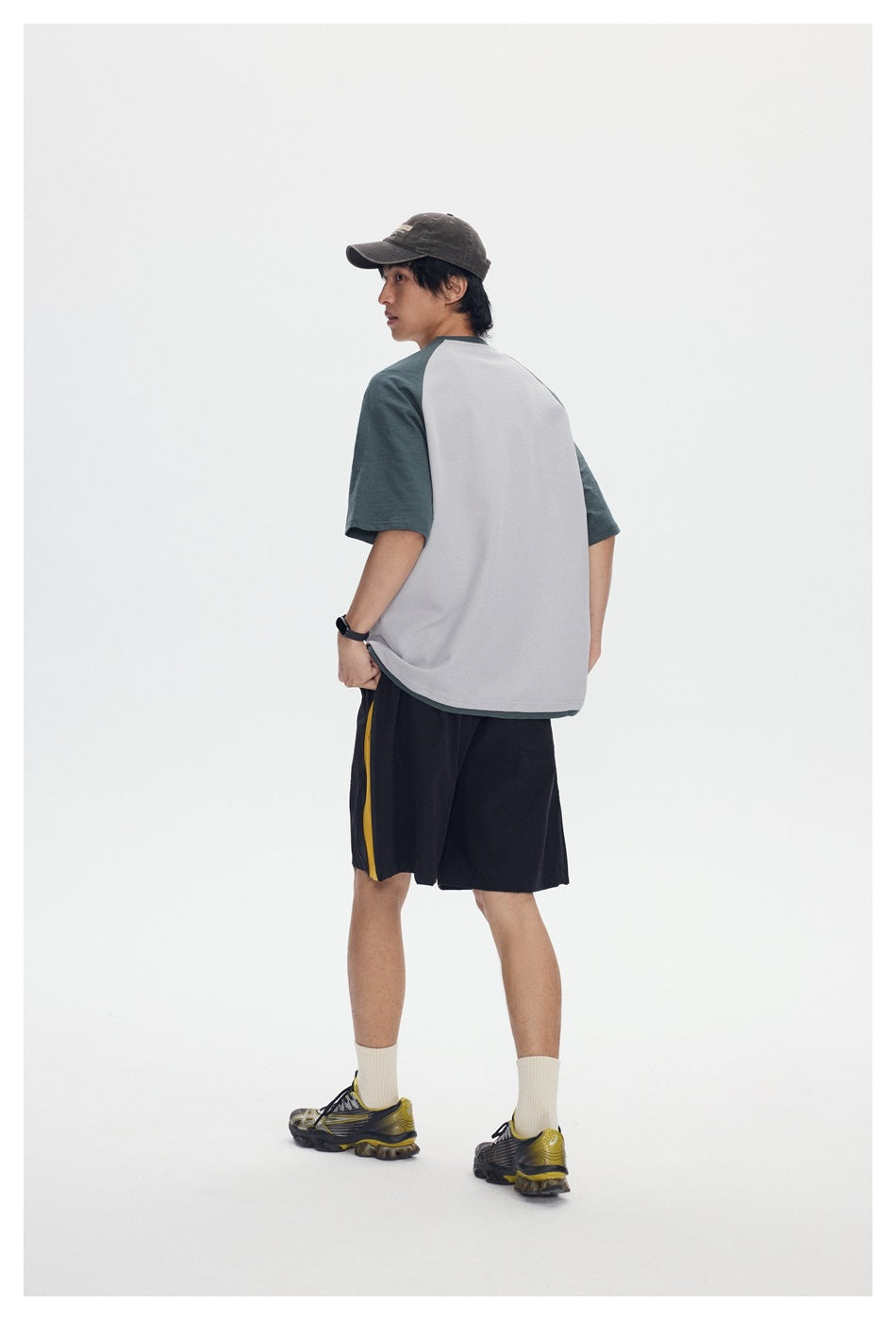 Back view of a model in a light gray T-shirt with green raglan sleeves, black shorts with a yellow stripe, cap and sneakers