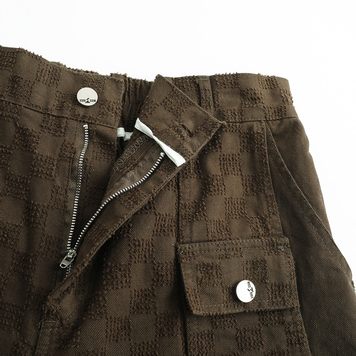 Close-up of brown checker-textured shorts showing the waistband, open zipper fly, and flap pocket.
