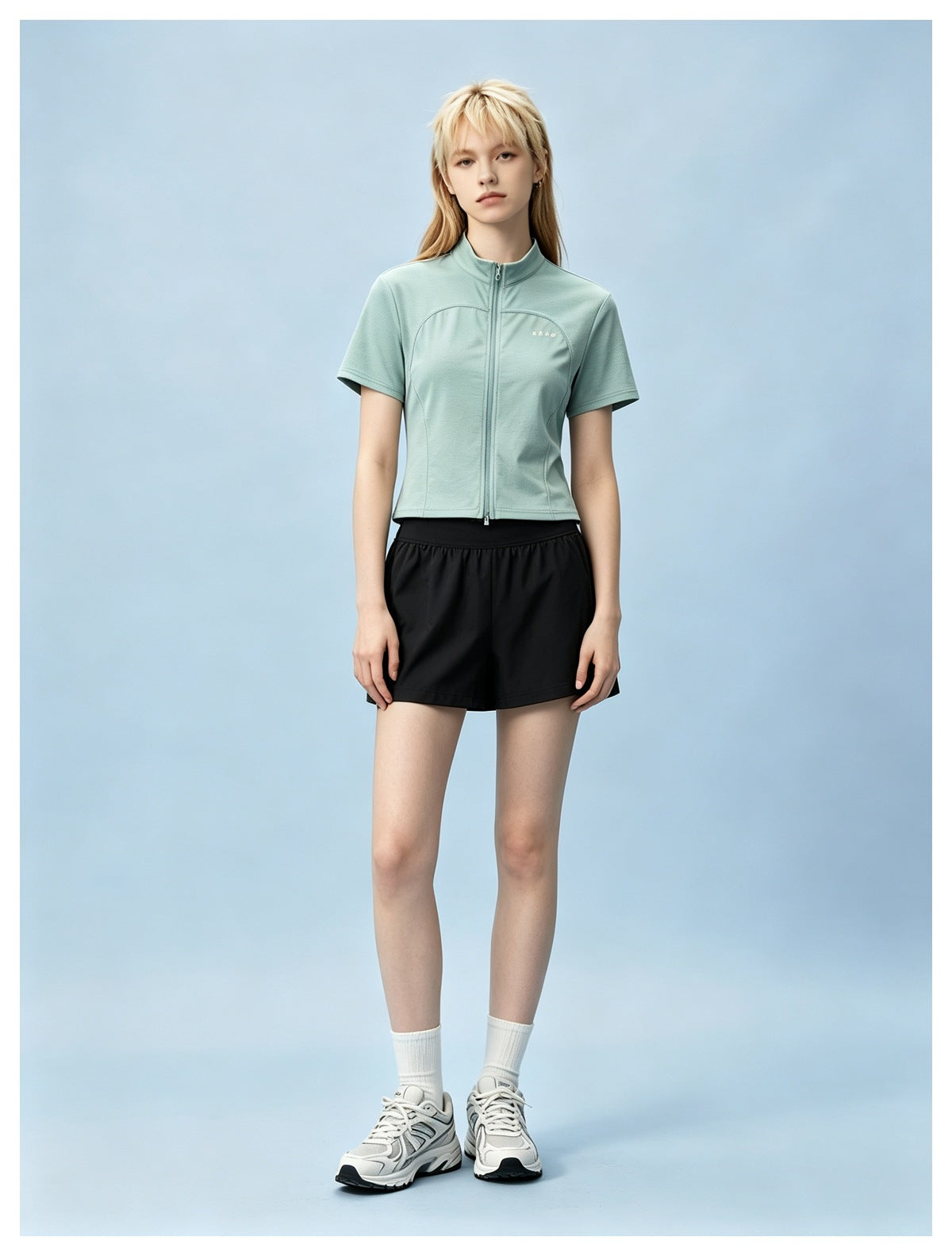 Model wearing a green short-sleeve zip-front top with black shorts and white sneakers against a blue backdrop