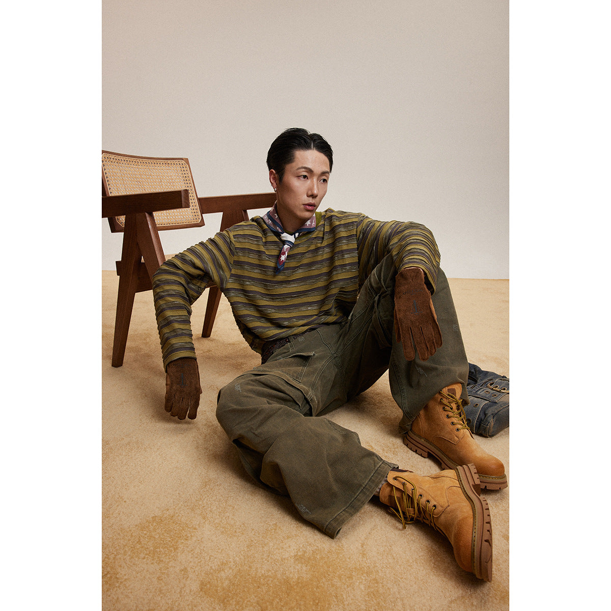 Seated model wearing an olive and brown striped long-sleeve shirt, olive cargo pants, gloves, and tan boots.