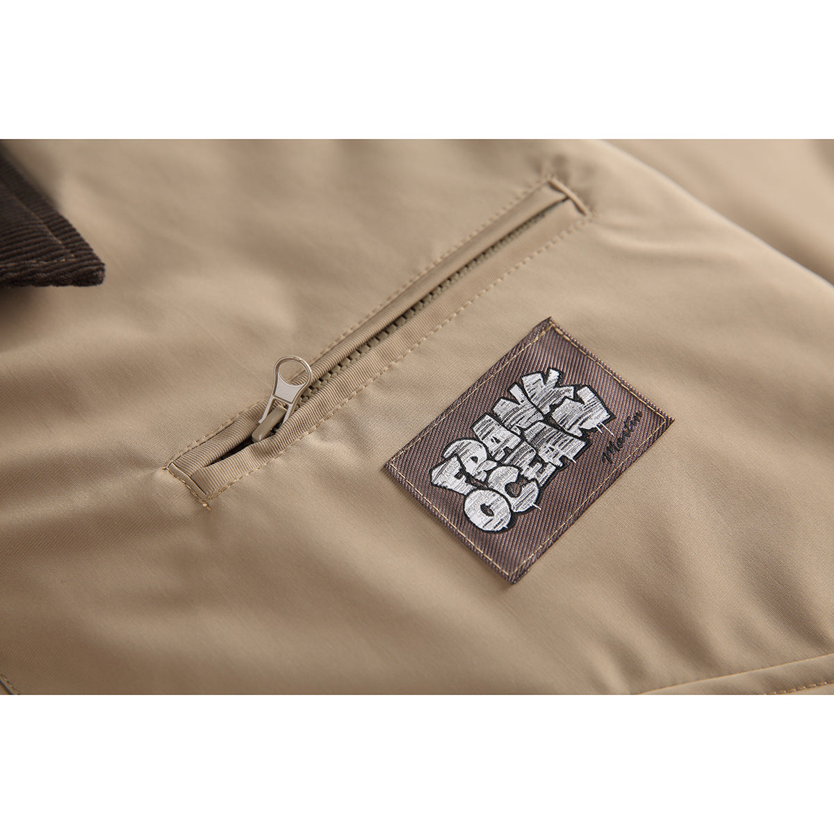 Close-up of a tan jacket's diagonal zip pocket and embroidered patch.