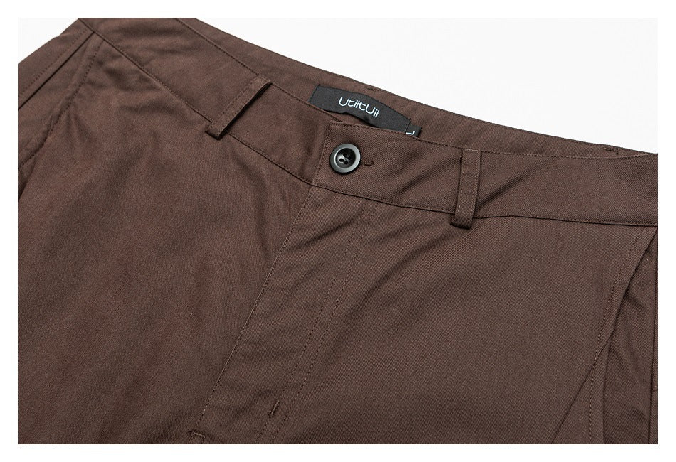 Close-up of dark brown trousers showing the waistband, button, belt loops, fly stitching, and a sewn-in label.