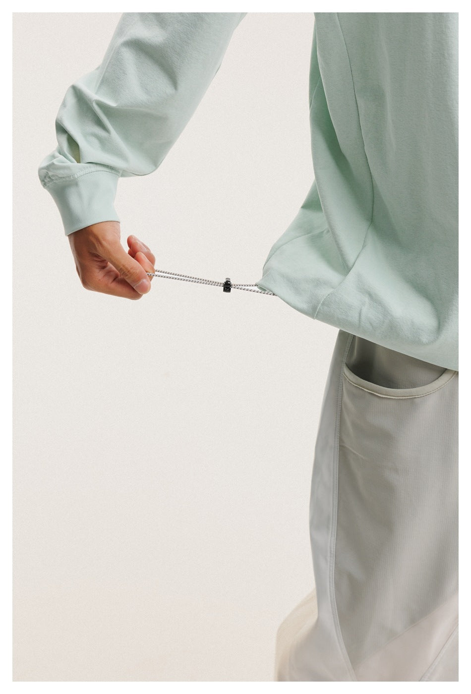 Close-up of a hand pulling the adjustable drawcord at the hem of a pale mint long-sleeve shirt.