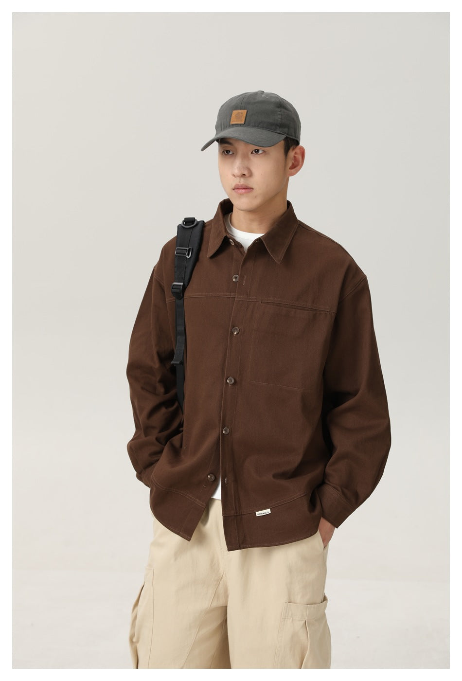 Model wearing an oversized dark brown button-up shirt with a chest pocket, cream trousers, and a gray cap.