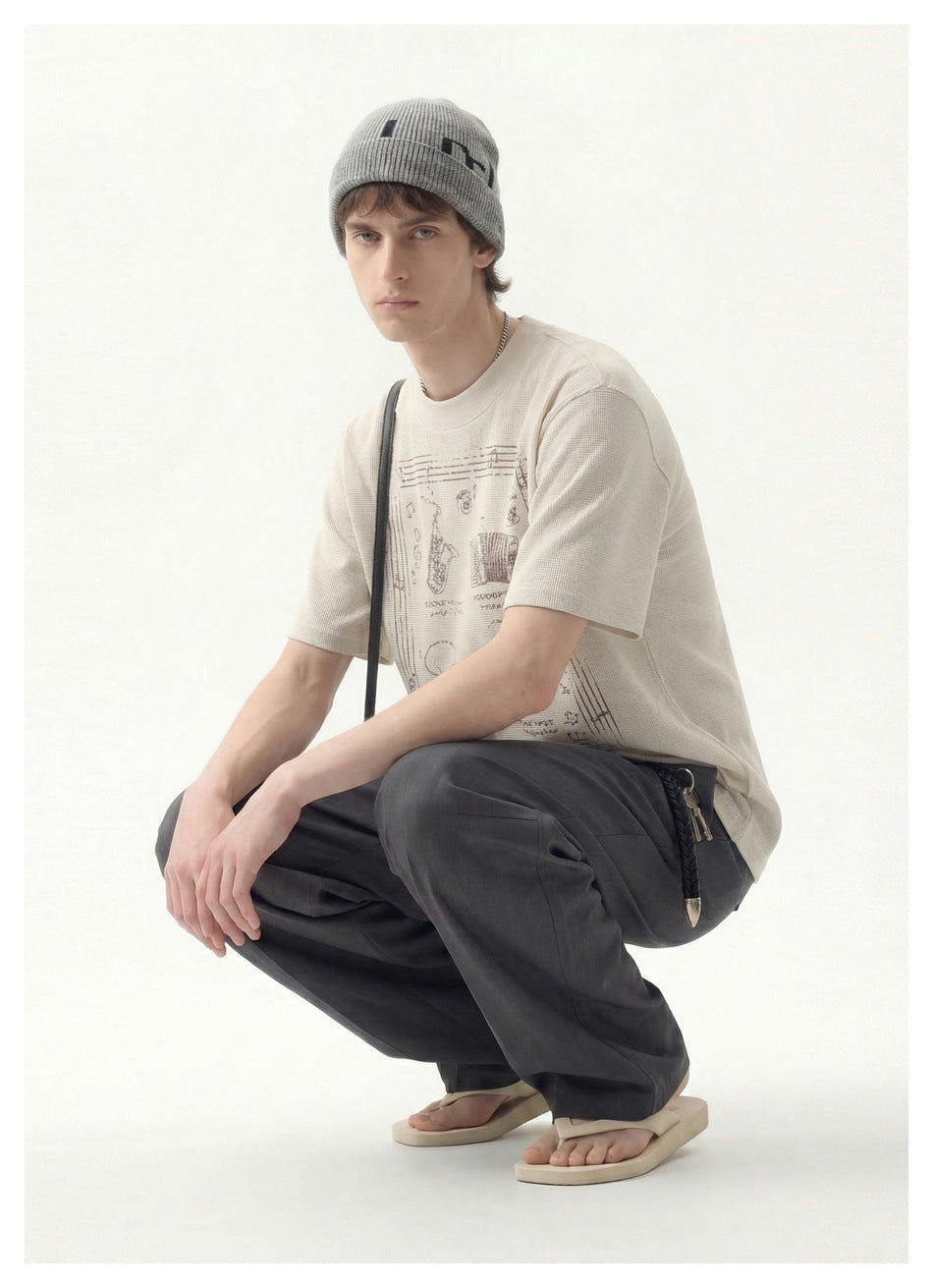 Crouching model wearing a cream graphic T-shirt, gray knit beanie, charcoal wide-leg trousers and sandals.
