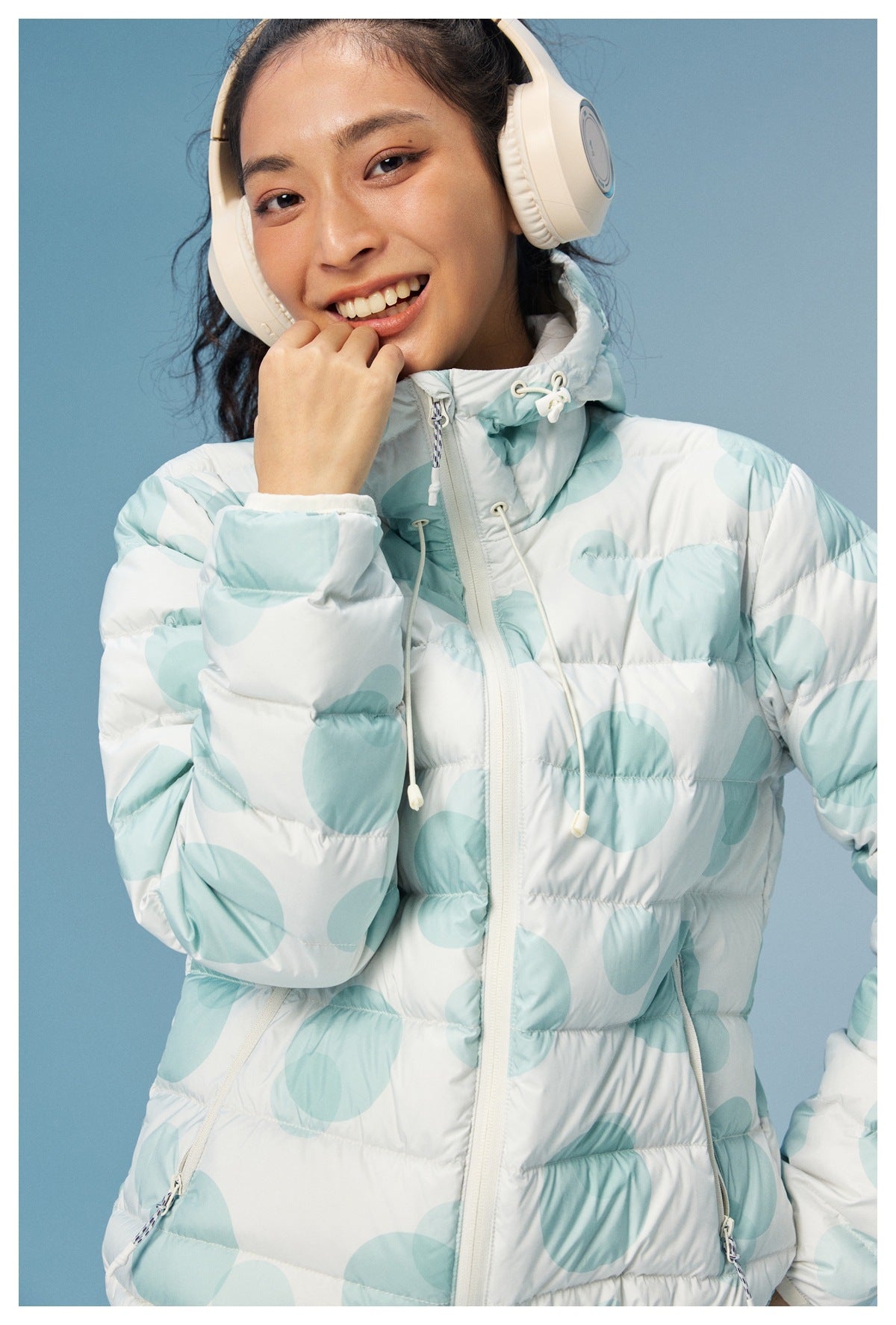 Close-up of a model wearing a white puffer jacket with pale aqua circles, showing the hood drawcords and front zipper.