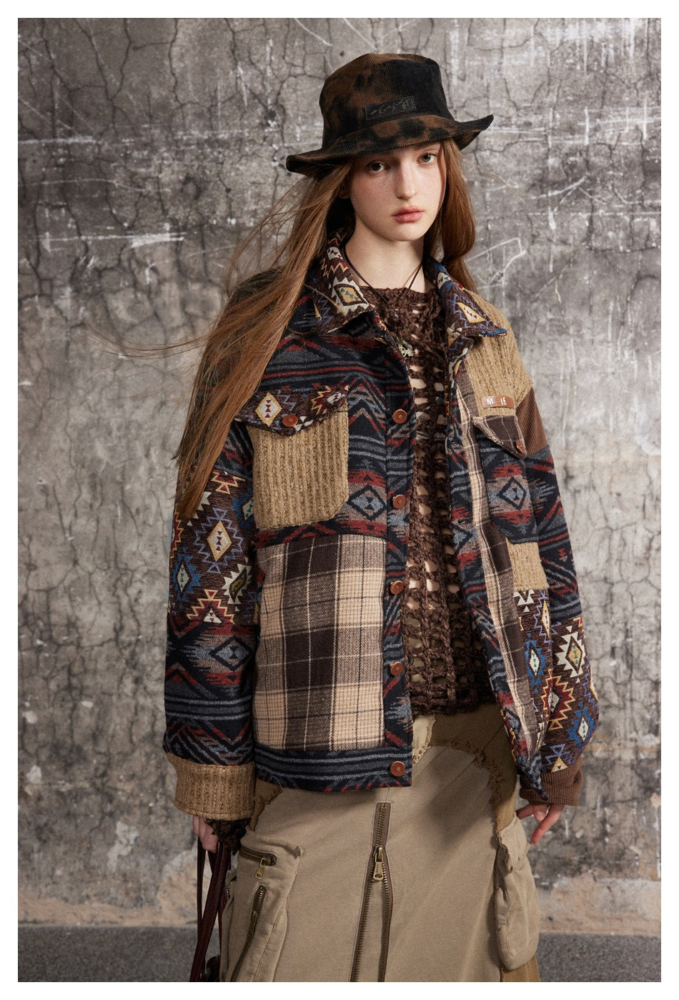 Model wearing a patchwork jacket with geometric and plaid panels, tan accents, and a dark wide-brim hat.