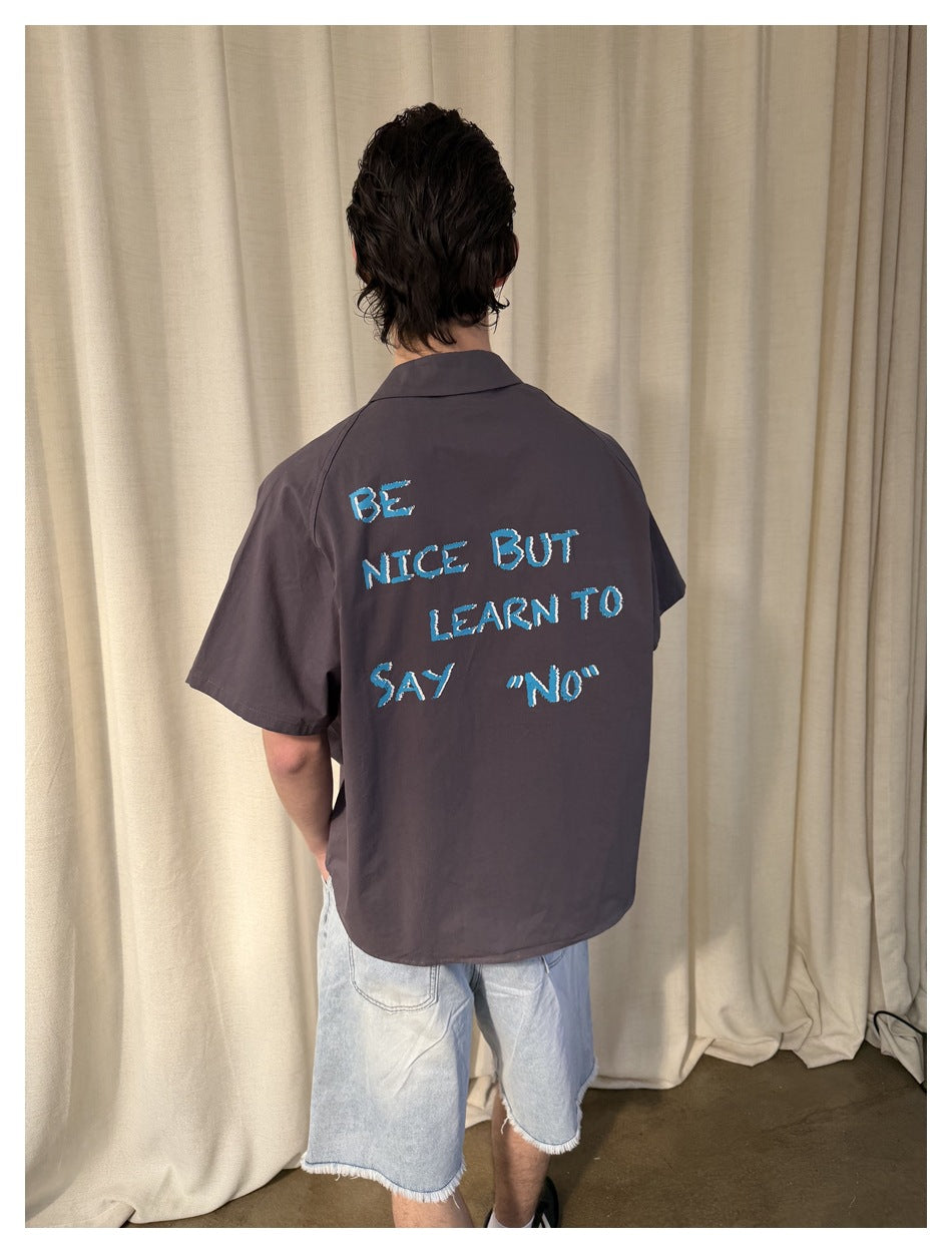 Close rear view of a charcoal shirt with blue text reading “BE NICE BUT LEARN TO SAY ‘NO’.”