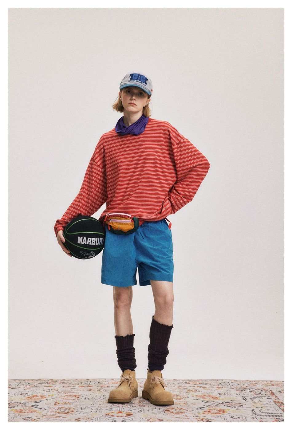 Person wearing an oversized red striped long-sleeve top with blue shorts, cap, tall socks and boots, holding a basketball