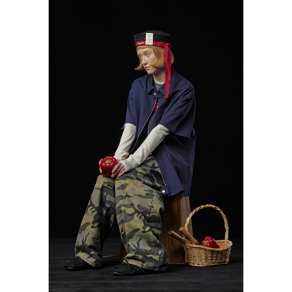 Seated model wearing an oversized navy short-sleeve zip shirt over layered tops and loose camouflage-print trousers.