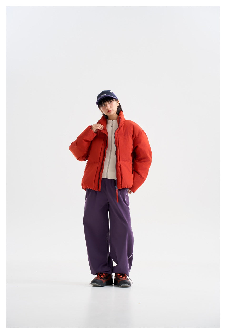 Model wearing a bright red-orange puffer jacket with a high collar, purple trousers, and a navy and purple cap.
