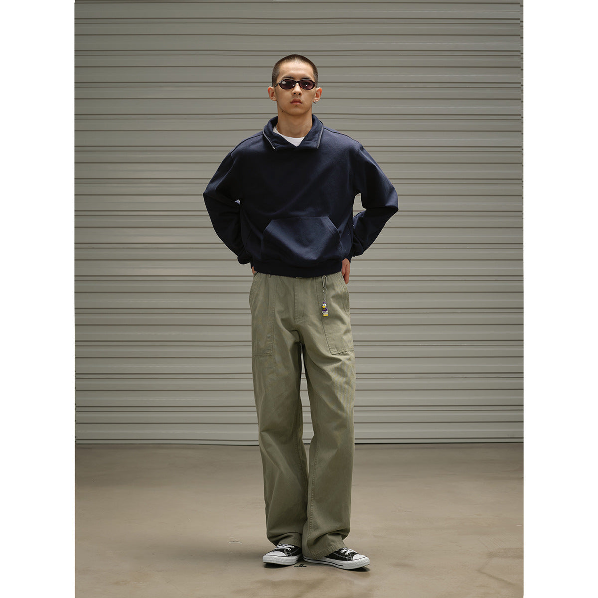 Model wearing a dark navy pullover with a high collar and kangaroo pocket, paired with loose olive trousers.