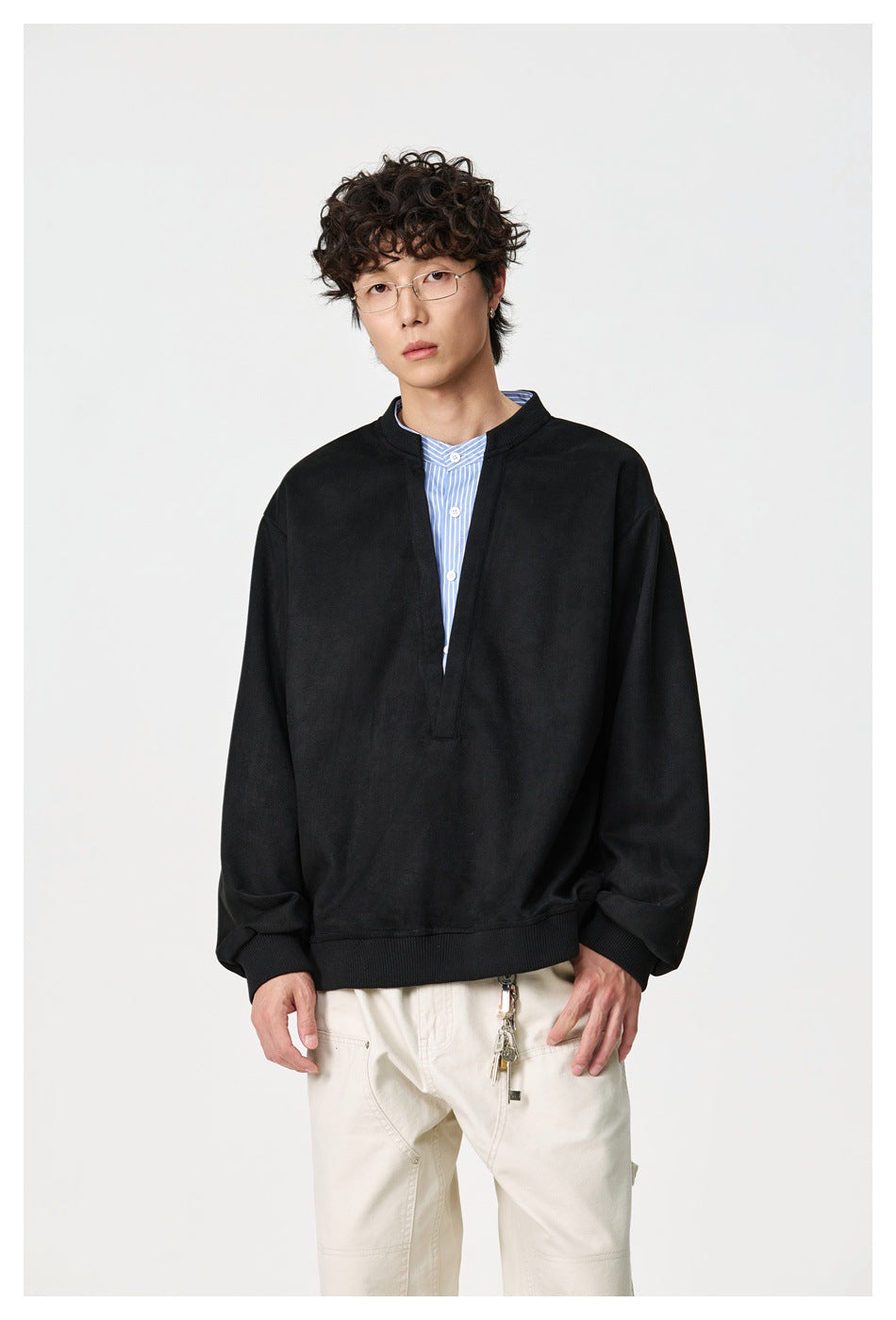 Front view of a model wearing a black oversized split-neck pullover over a blue striped shirt and cream trousers.