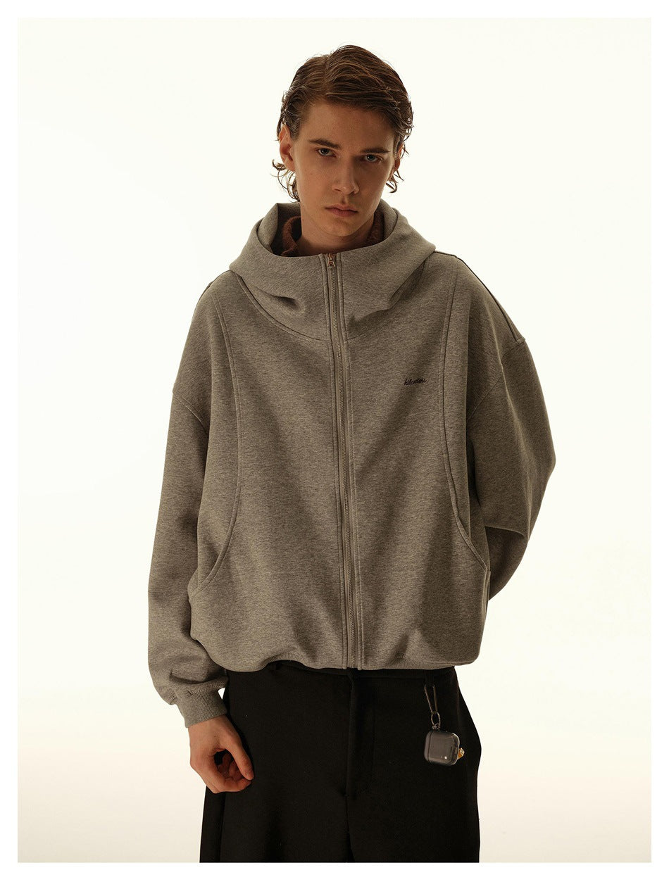 Model wearing a gray zip-up hoodie with high hood and curved seams over black trousers, front view