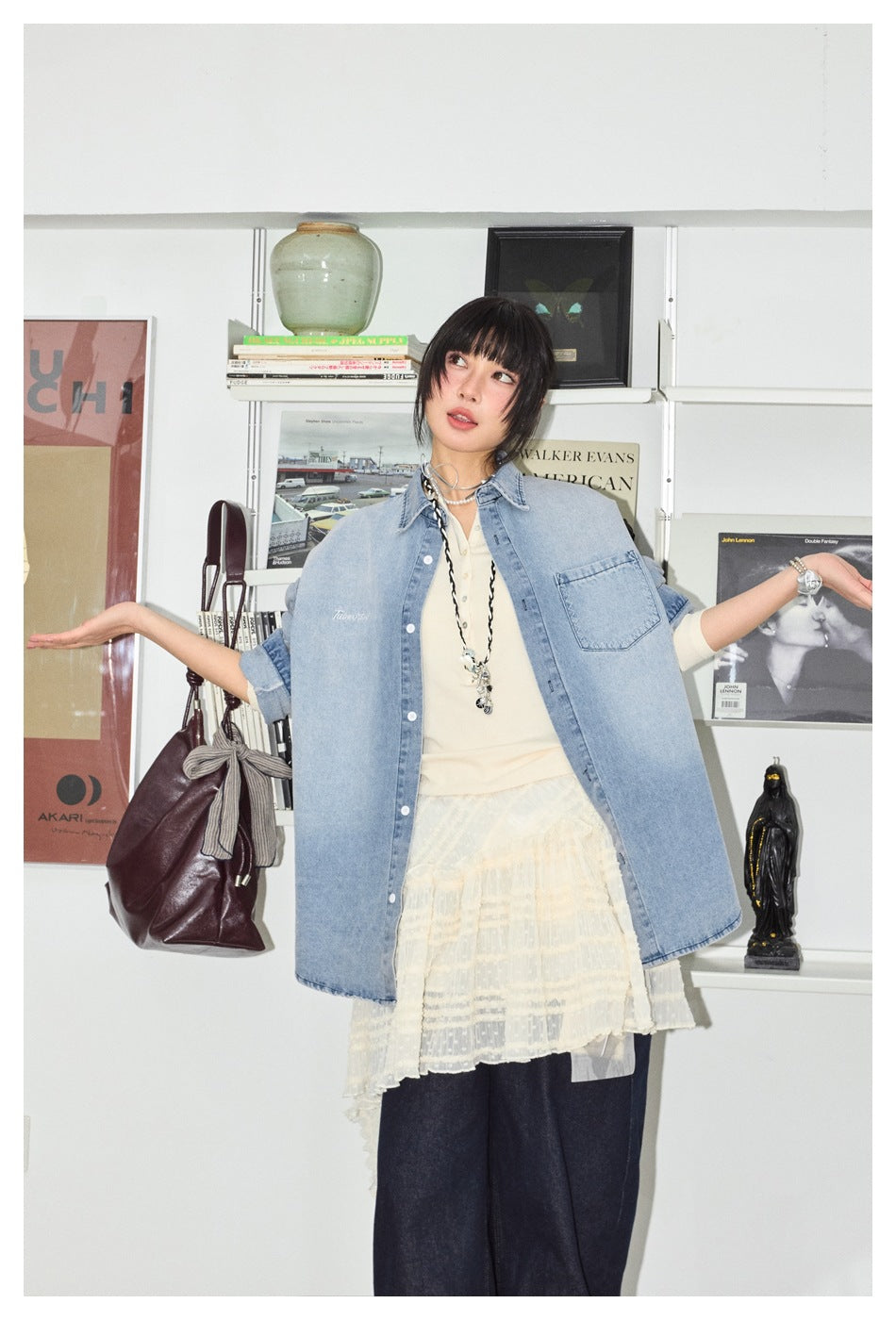 Model wearing an oversized light blue denim shirt with a chest pocket, open over a cream top and lace skirt.