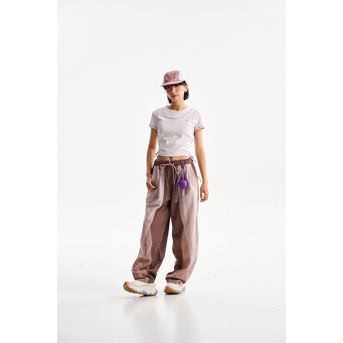 Model wearing a pale lavender cropped tee, loose taupe trousers and a pink cap.