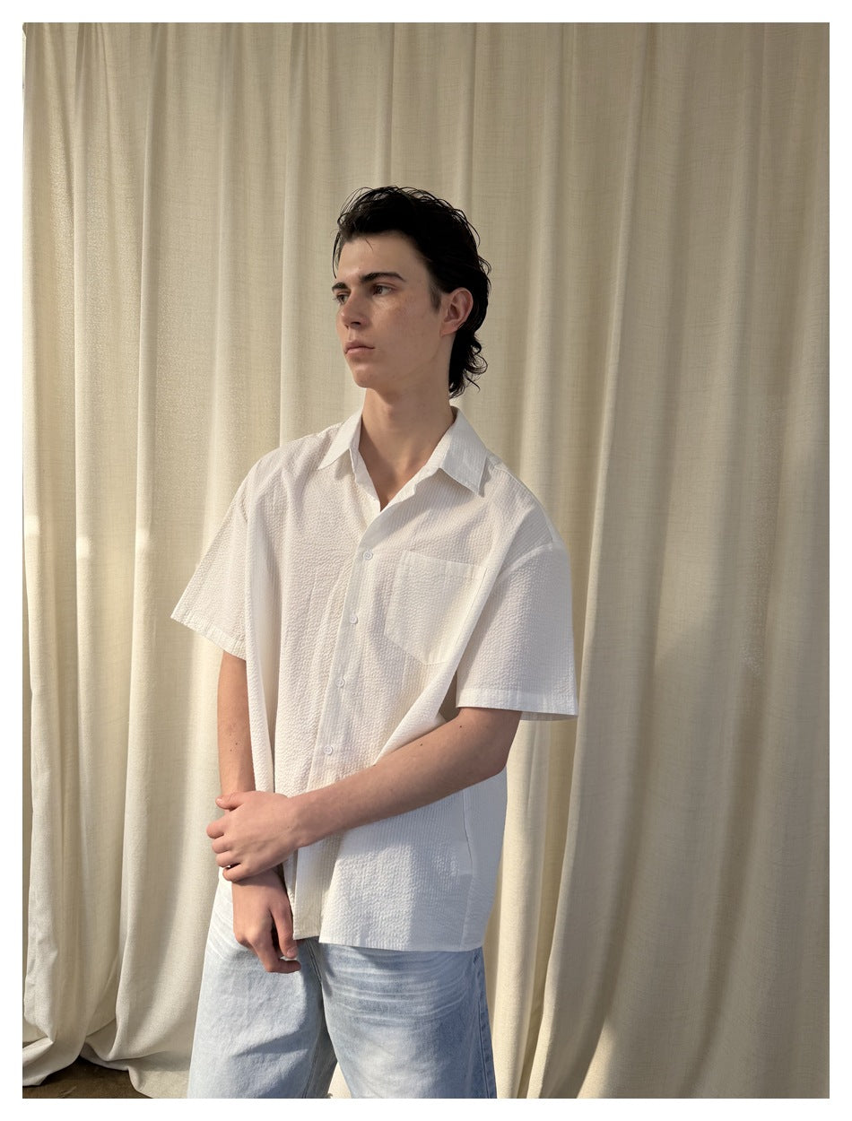 Model wearing a white textured short-sleeve button-up shirt with chest pocket and light jeans, against a cream curtain