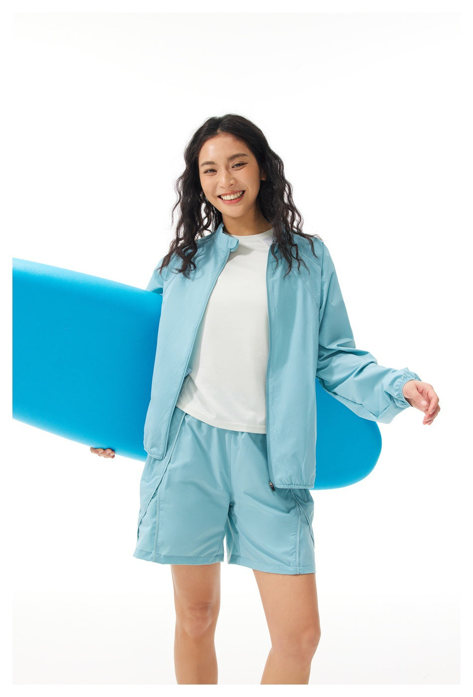 Model wearing a light blue zip jacket and matching shorts over a white shirt, holding a blue board.