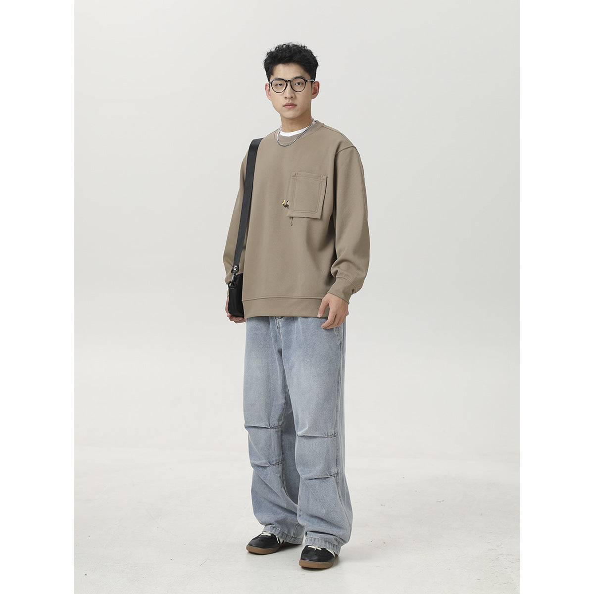 Man wearing a taupe crewneck sweatshirt with a large chest pocket and loose light-wash jeans, shown from the front.