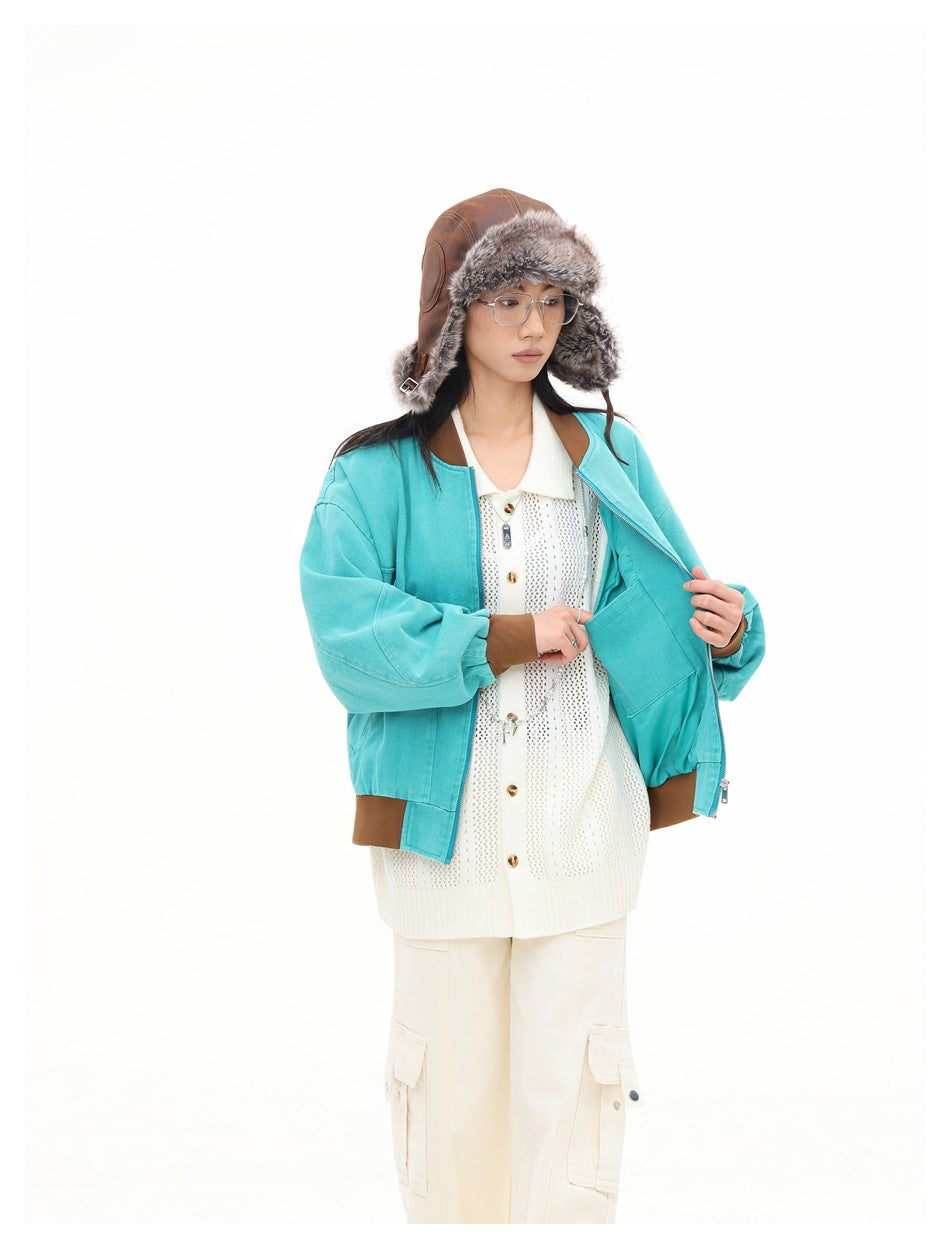 Model holds open a turquoise bomber jacket, showing its brown trim and lining over a cream knit cardigan.