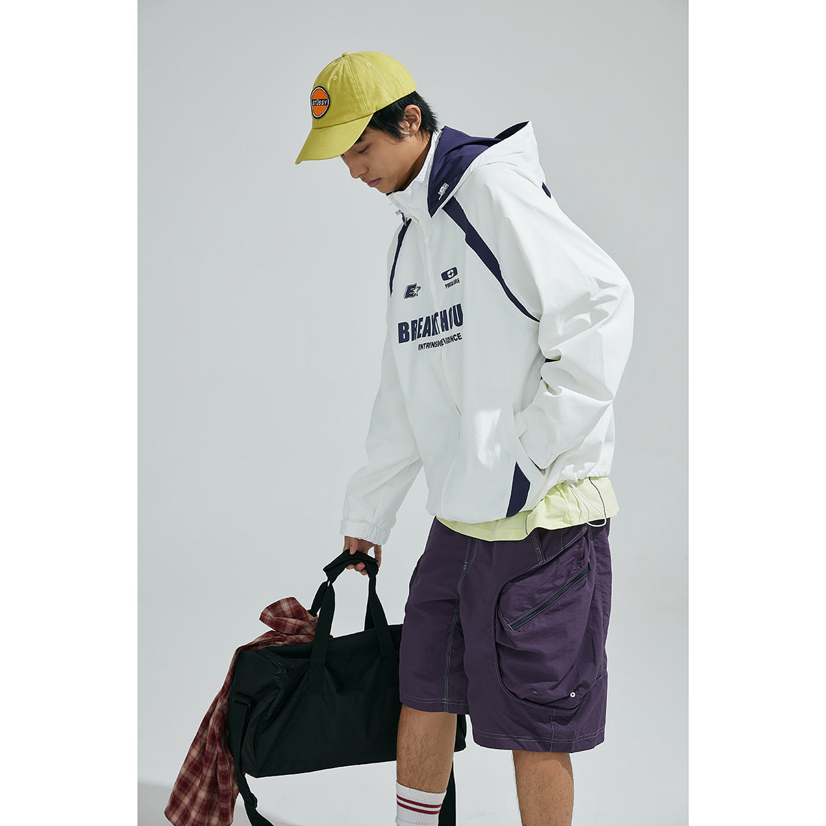 Model in a white hooded jacket with navy accents and chest graphics, purple shorts, and a yellow cap.