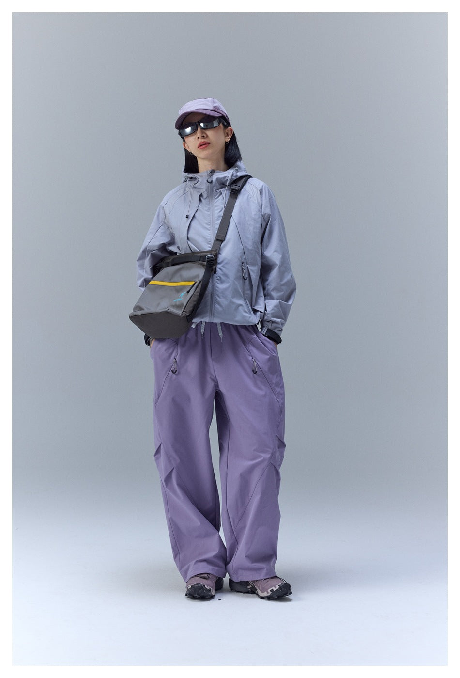 Model wearing a zipped light gray hooded jacket and loose lavender pants with a crossbody bag.