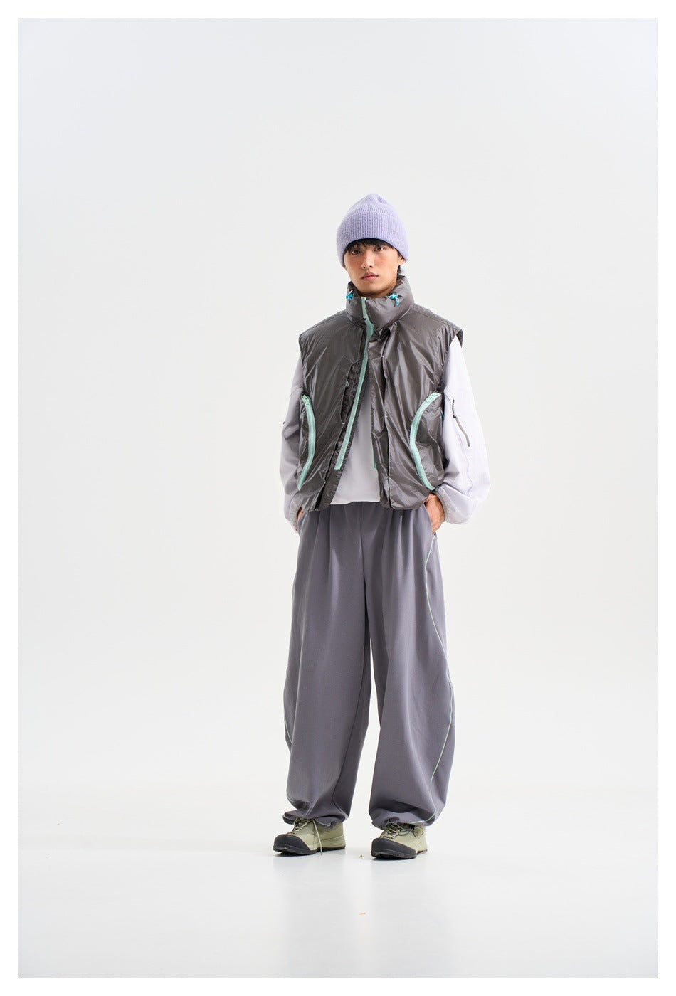 Model wearing a glossy charcoal sleeveless vest with aqua trim, white sleeves, loose gray trousers and a lavender beanie.