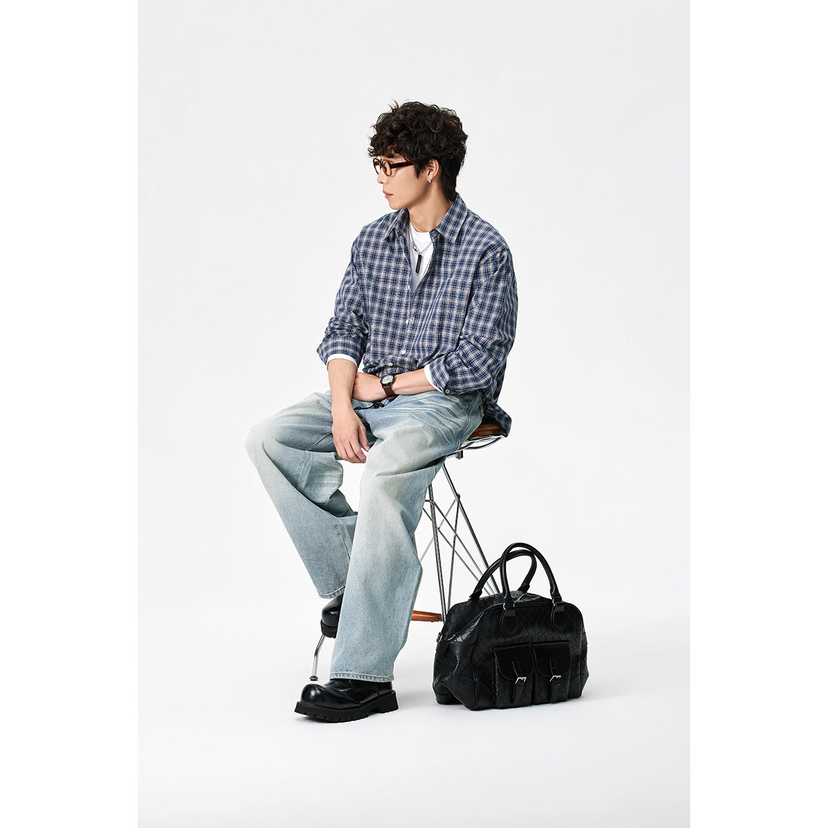 Model seated in a blue plaid button-up shirt over a white tee, with loose light-wash jeans and black boots.