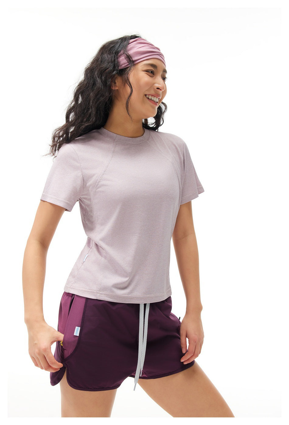 Three-quarter view of a woman wearing a pale lilac short-sleeve shirt and plum running shorts.