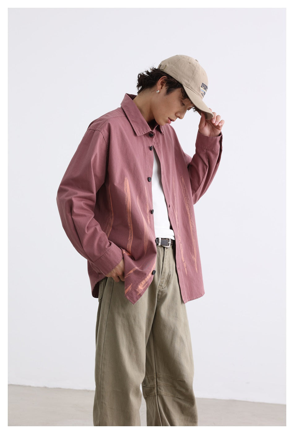 Model wearing a dusty rose oversized button-up shirt with tan brushstroke graphics, khaki trousers, and a beige cap.