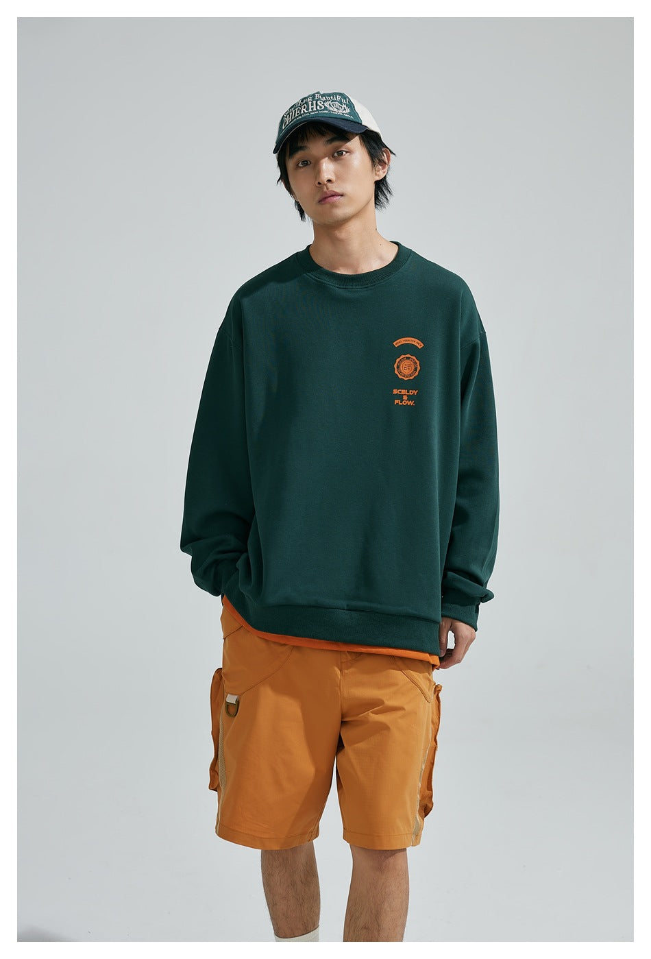 Model wearing a dark green sweatshirt with a small orange chest emblem and ochre cargo shorts, shown from the front.