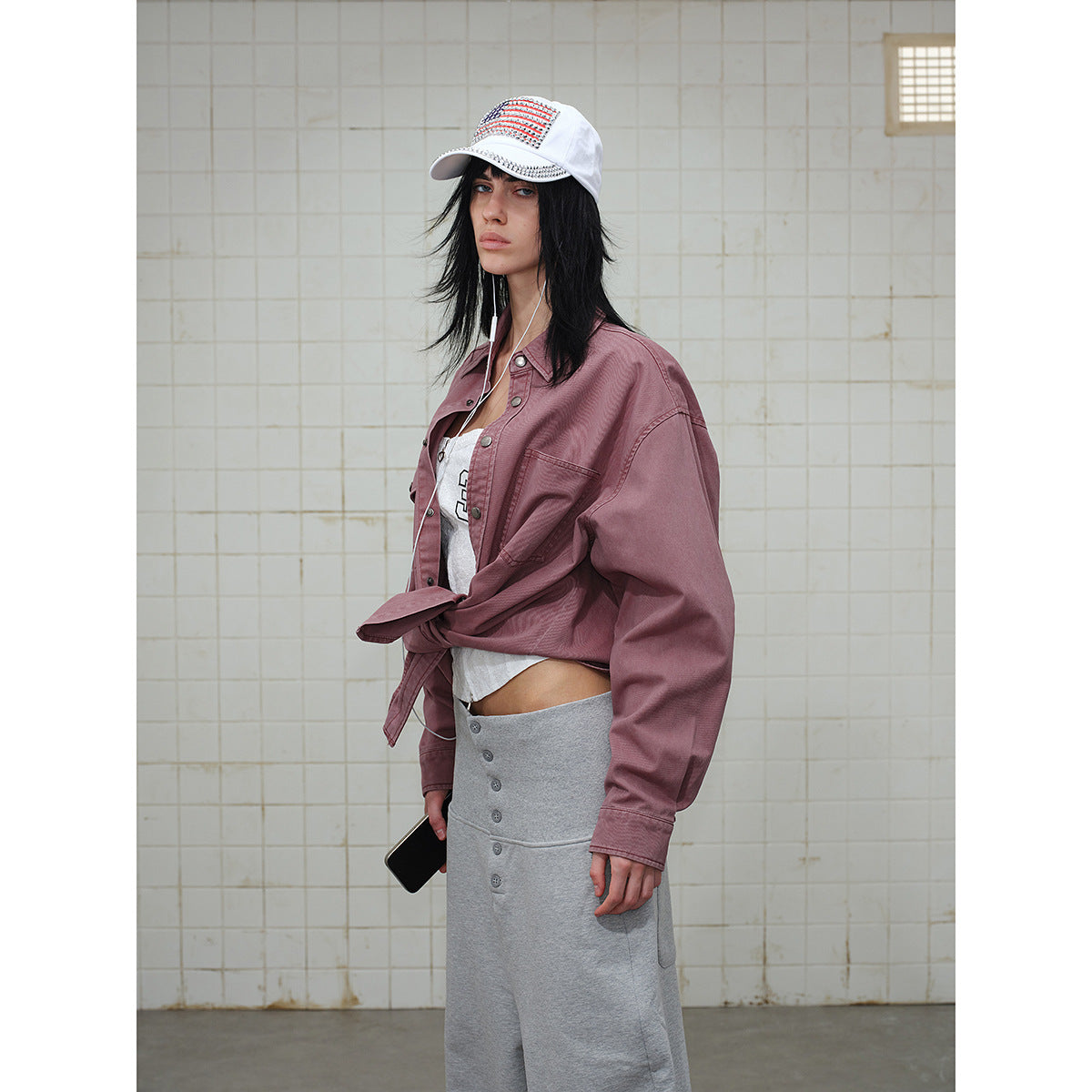 Side view of a model in a dusty mauve shirt tied at the waist, gray pants, and a white cap.