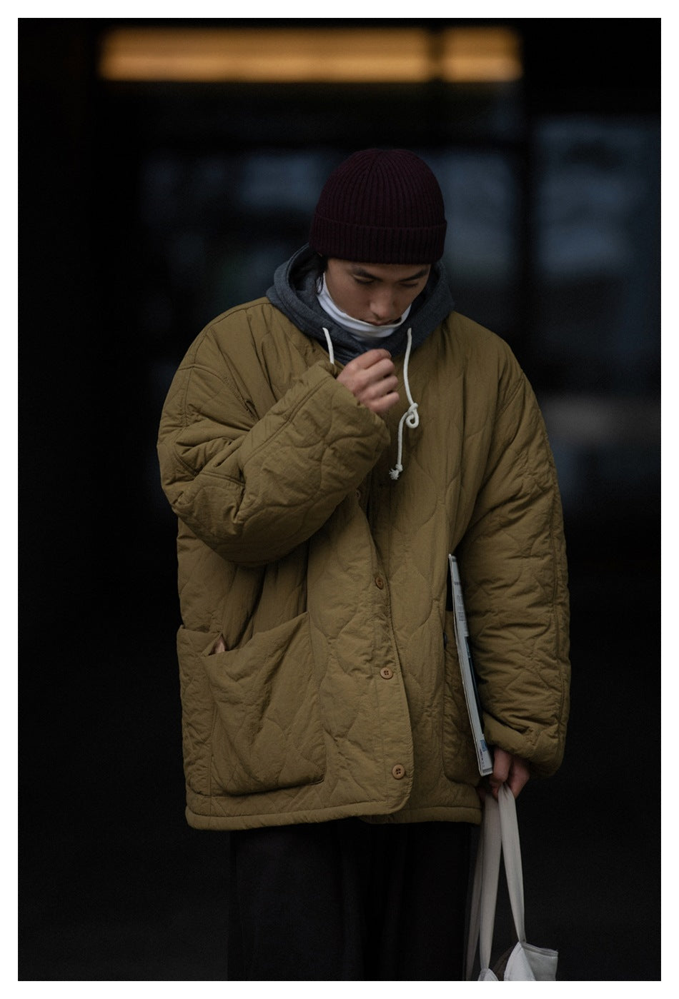 Closer front view of an olive quilted jacket with button closure and patch pockets, worn over a gray hoodie.
