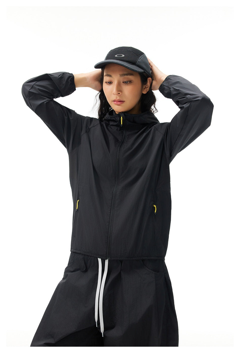 Model wearing a black hooded zip-up jacket with yellow zipper pulls, black trousers, and a black cap.