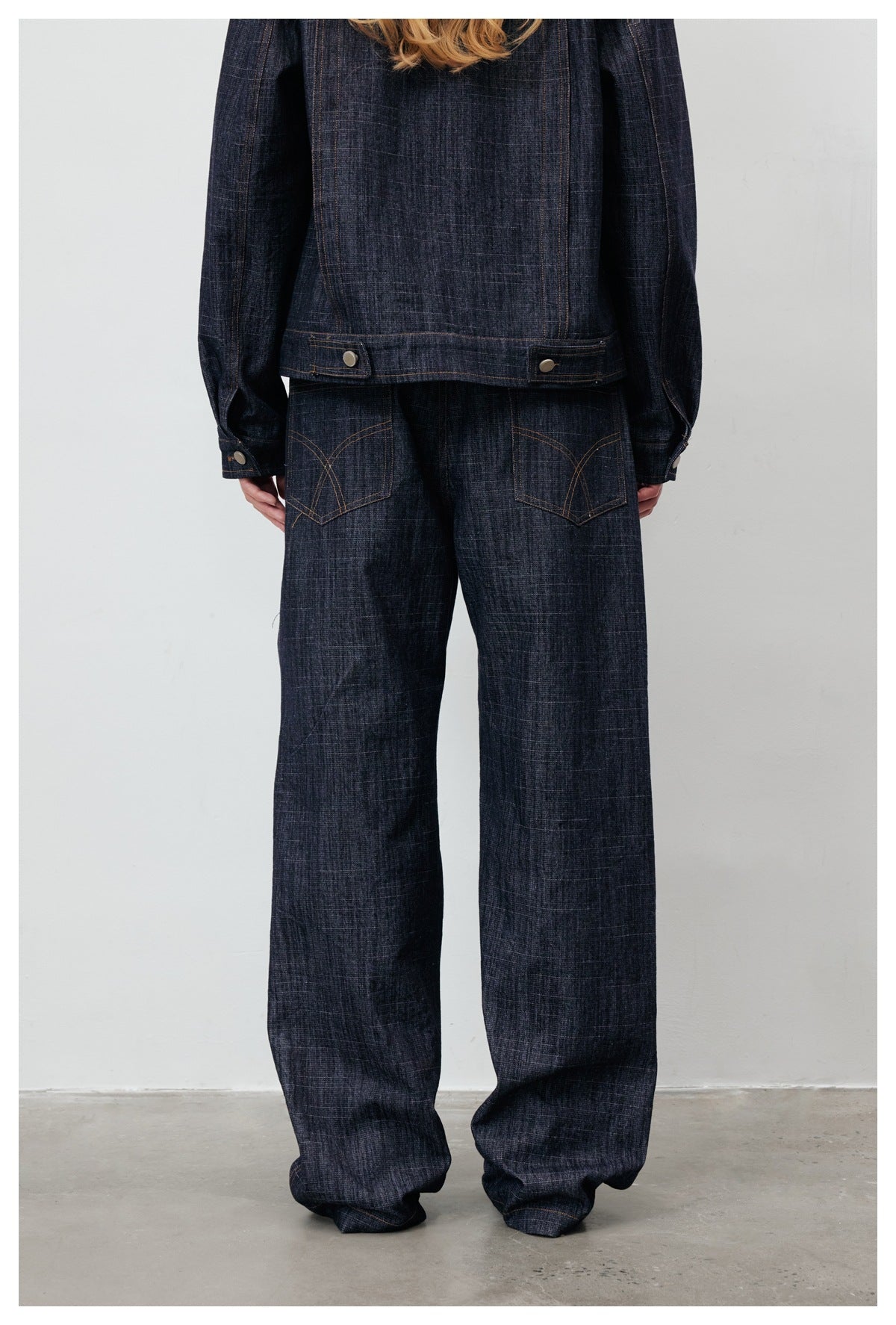 Rear view of a model wearing a dark indigo denim jacket and matching loose wide-leg jeans with curved-stitched pockets.
