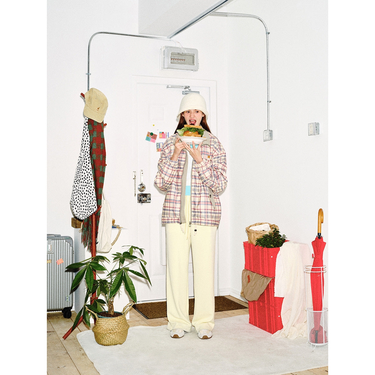 Model wearing a pastel plaid zip jacket and cream wide-leg pants, holding a plate of food.