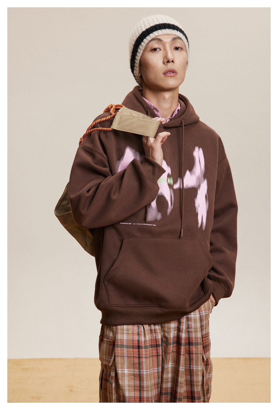Model wearing a dark brown graphic hoodie with plaid trousers and a tan shoulder bag.