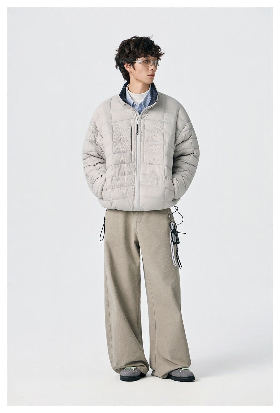 Model wearing a light gray quilted zip jacket and loose beige trousers, standing with hands in pockets.