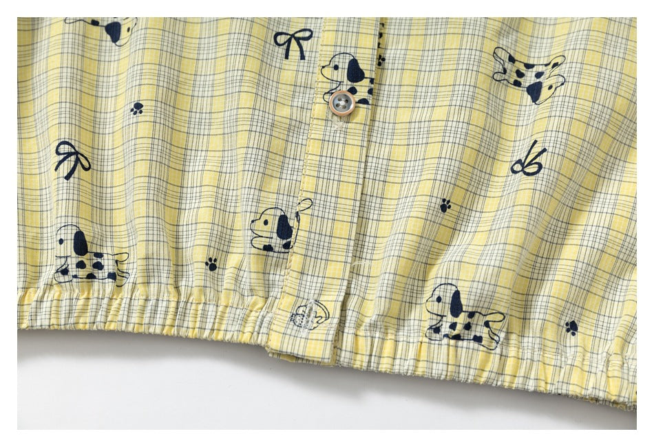 Close-up of a yellow plaid garment with navy dog and bow prints, a button placket, and a gathered hem.