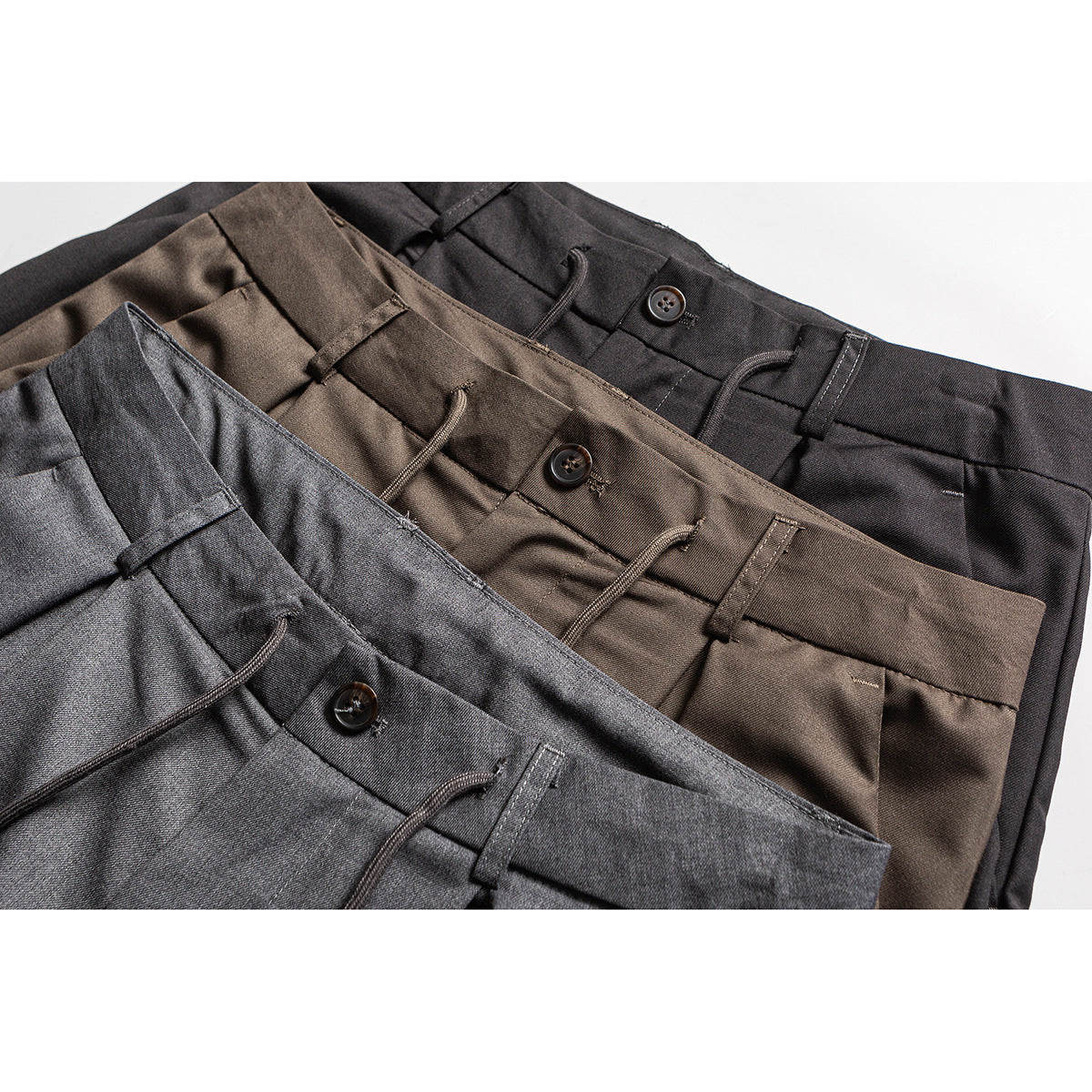 Close-up of three trousers in charcoal gray, brown, and black, showing waistbands, buttons, belt loops, and drawstrings.