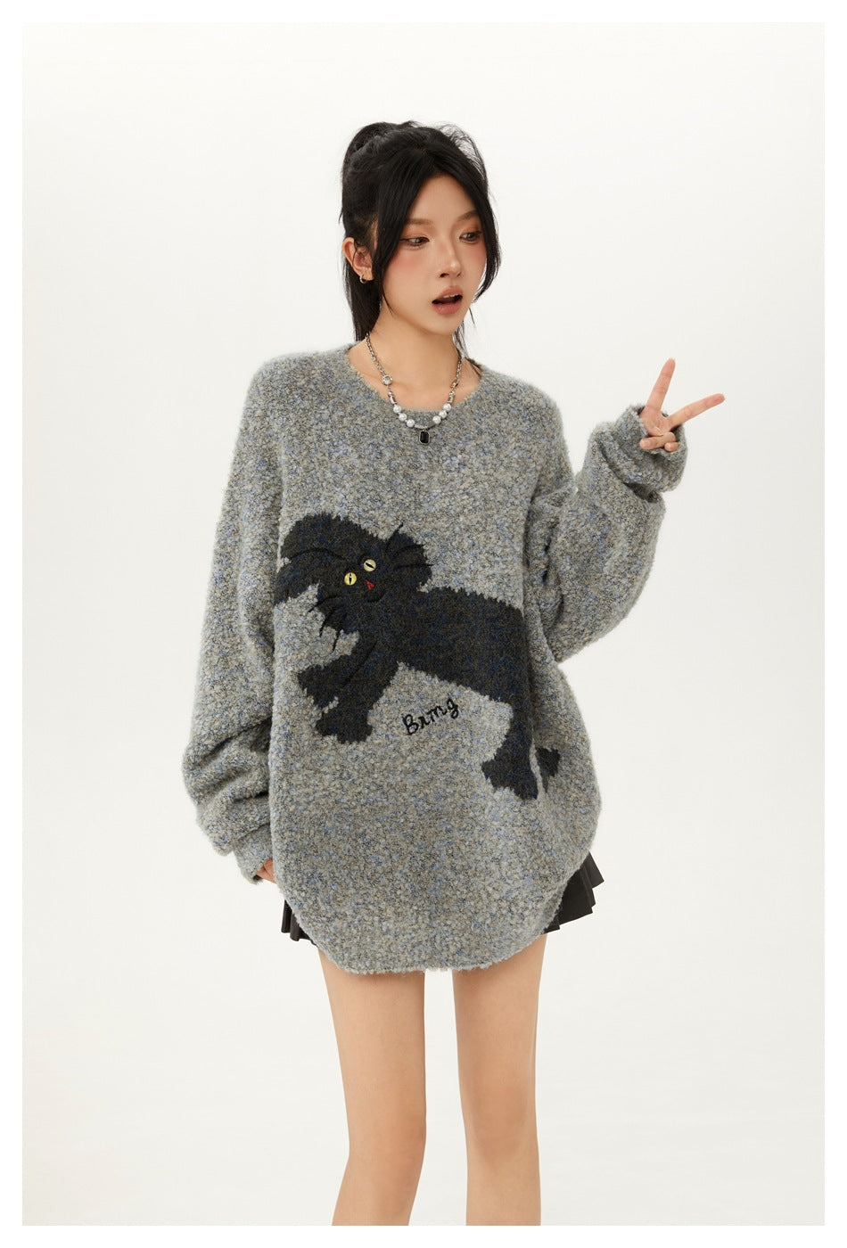 Model wearing an oversized gray-blue sweater with a large black cat graphic and raising a peace sign.