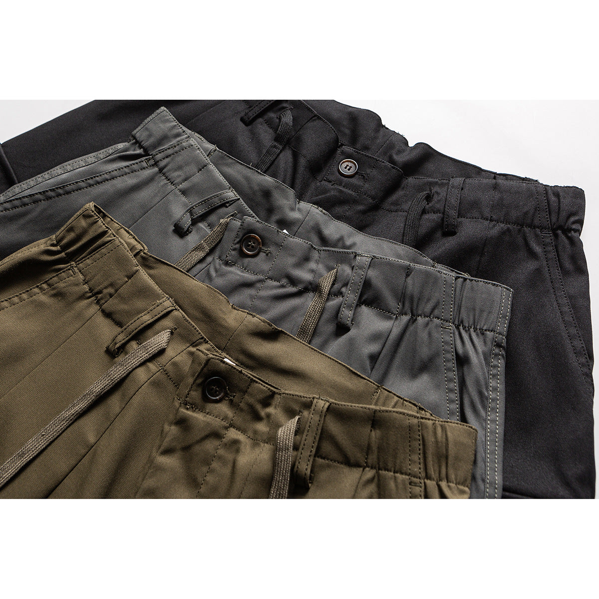 Close-up of overlapping olive, gray, and black pants waistbands with buttons, belt loops, and drawstrings.