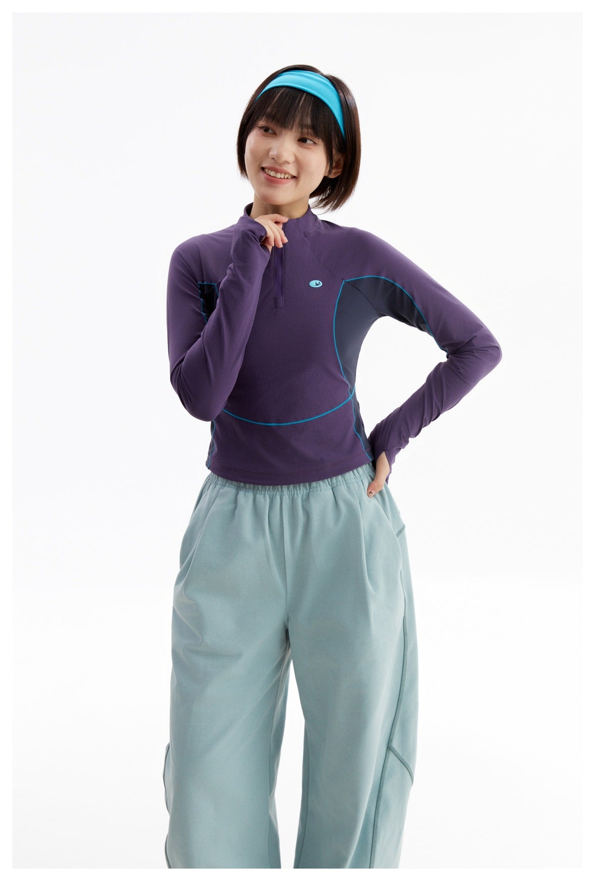 Model wearing a purple quarter-zip top with dark contrast panels and teal piping, paired with pale blue trousers.