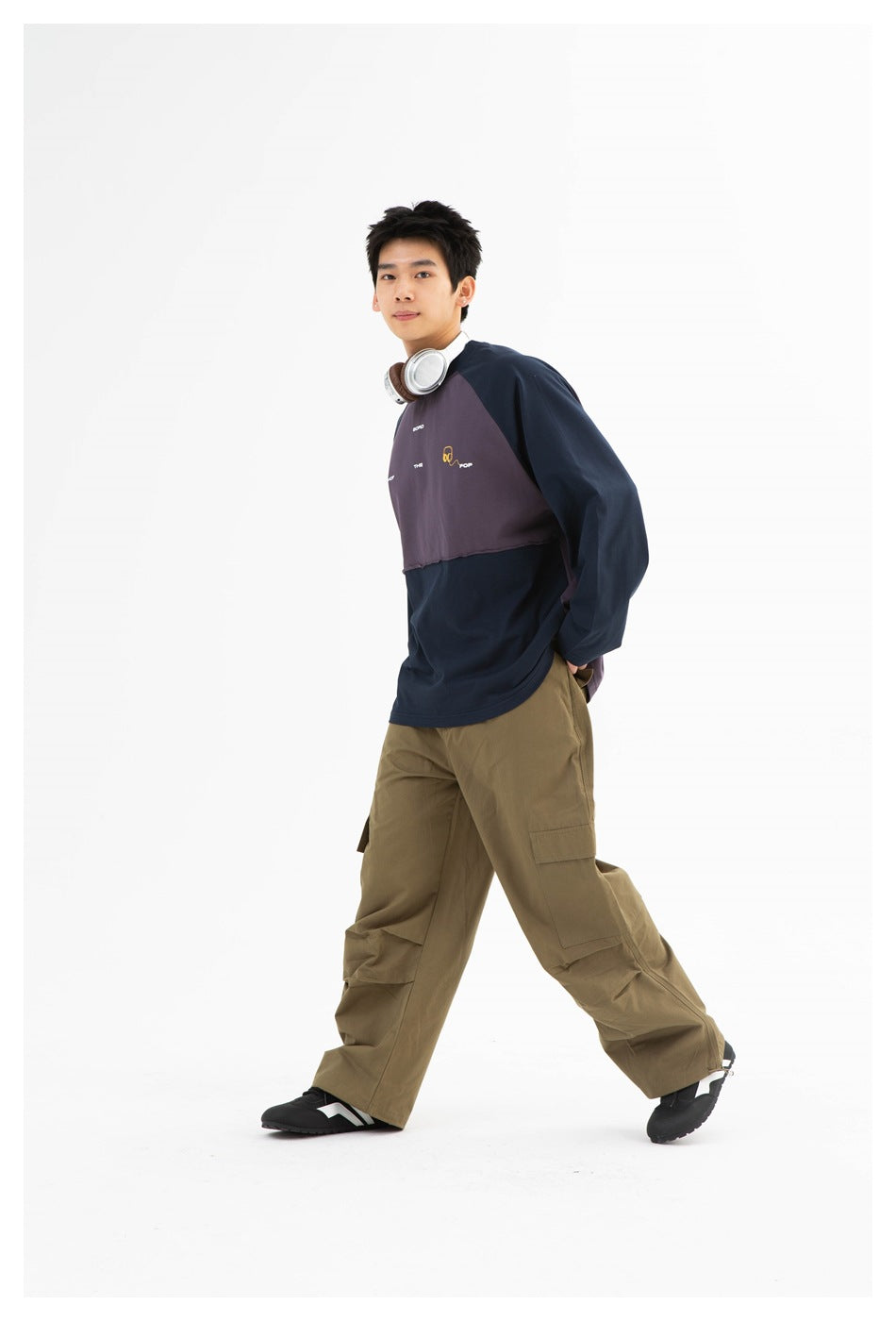 Model walking in navy and purple-gray color-block long-sleeve top with olive cargo pants and headphones around neck