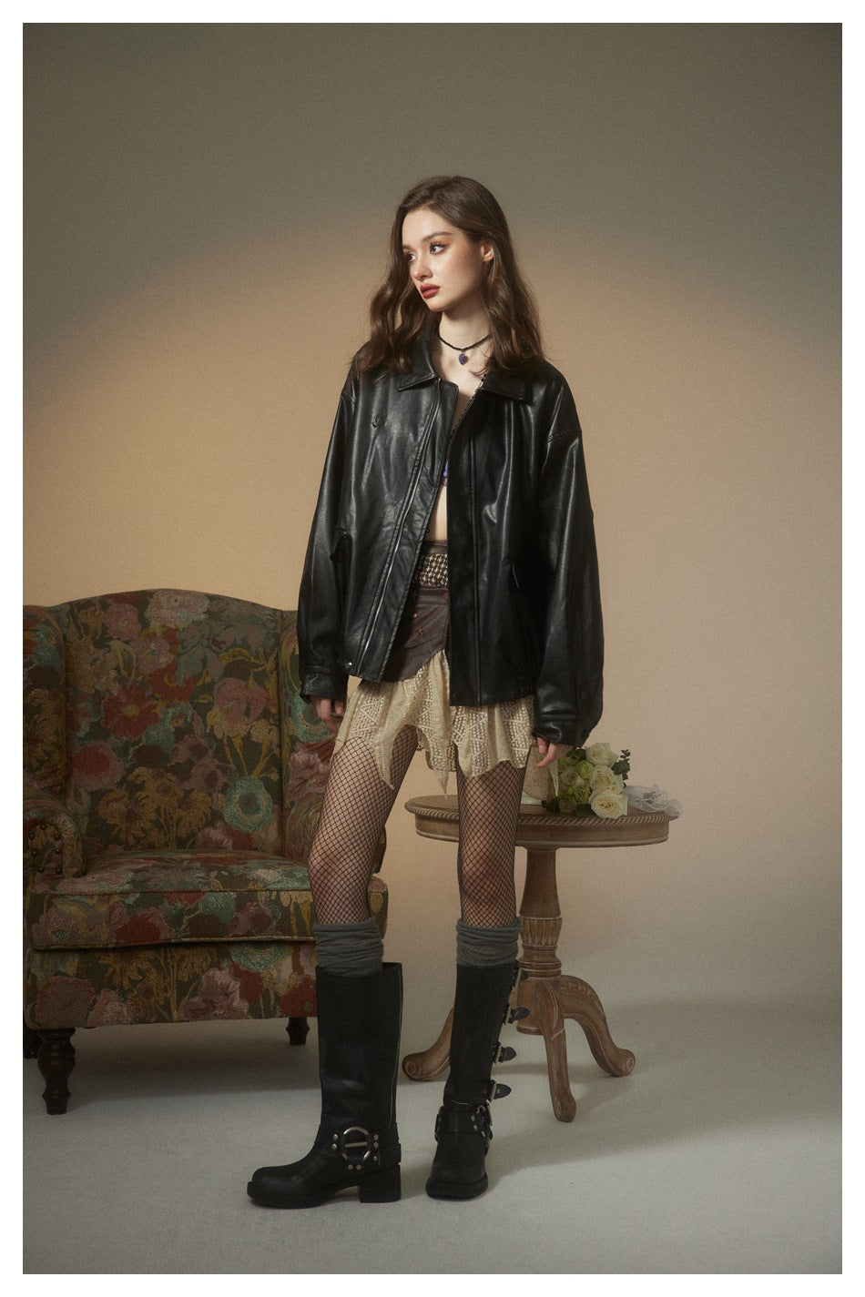Model in a glossy black oversized jacket with a pointed collar and zip front, shown with a cream lace skirt and tall boots.