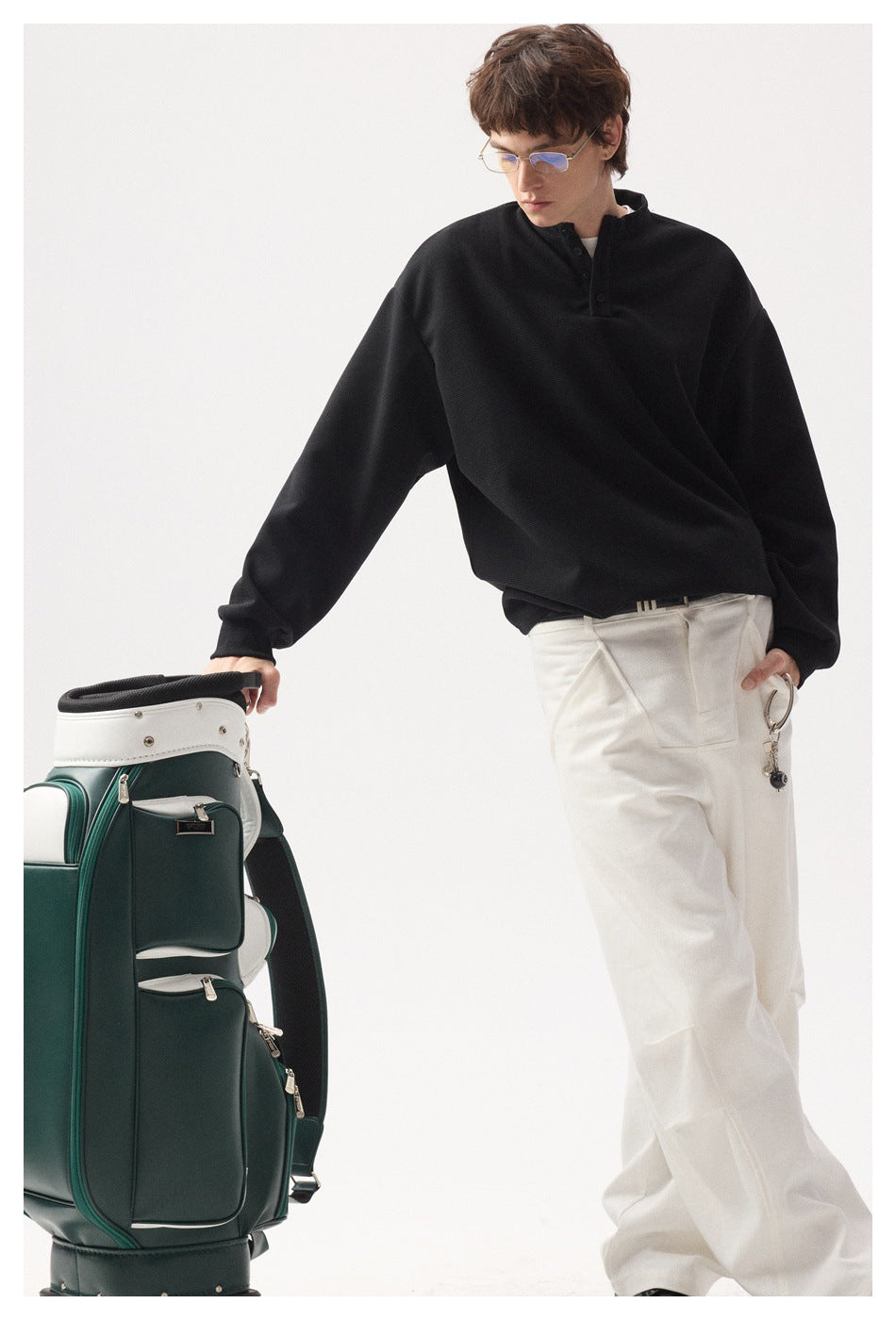 Model wearing an oversized black button-neck top and white trousers beside a green golf bag.