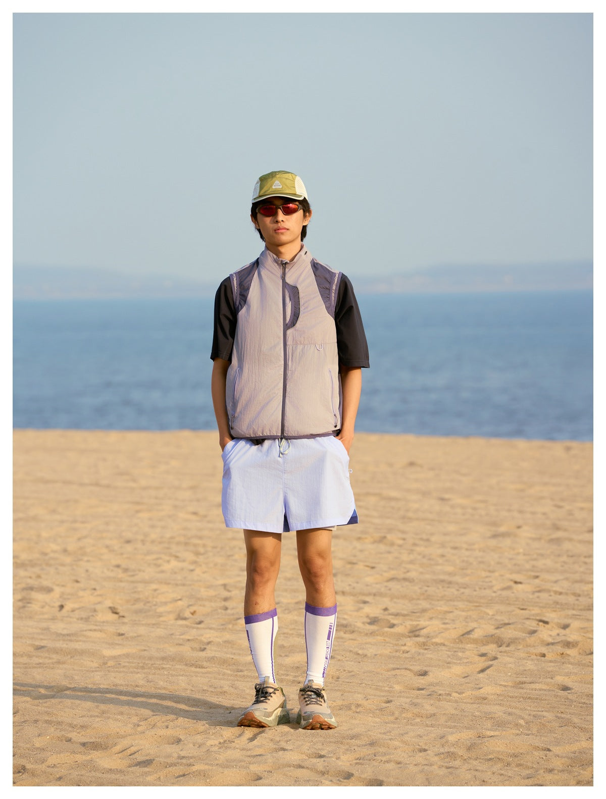 Person on a beach wearing a gray-lavender zip vest with purple panels over a dark tee, light blue shorts and a cap